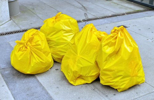 Four large yellow rubbish bags made of durable, slightly crinkled plastic are placed on a grey concrete pavement, with some bags leaning against one another in a casual arrangement. The bags appear filled with waste, as indicated by their bulky and rounded shape, with some slightly bulging at the top where they are gathered and tied. The pavement surface includes a drainage grate and subtle surface textures, typical of a paved outdoor area in a residential or commercial setting, possibly near a street or alleyway. The background shows part of a building or pavement edge, with a slight indentation or curb visible. This scene relates to rubbish collection or removal, which Business Waste Removal Muswell Hill services, ensuring proper disposal of waste materials.