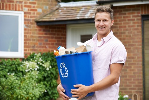 A young man with short, light brown hair and a friendly smile is standing outside a residential building in Muswell Hill, holding a blue recycling bin filled with various waste items, including cardboard, plastic bottles, and paper. He is dressed in a light pink button-up shirt and is positioned on a paved pathway beside a garden with green foliage and white flowers. Behind him, there is a brick house with a white-framed window and a sloped roof with brown tiles. The scene appears to be during daytime with natural lighting, and the background suggests a typical suburban environment in north London. Business Waste Removal Muswell Hill offers rubbish collection and disposal services in this area, helping local residents manage their household waste efficiently.