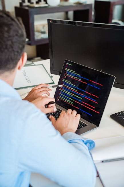 A person in a light blue shirt is seated at a desk, working on a laptop that displays lines of colorful computer code on its screen. The individual's right hand is holding a pen or stylus, pointing to